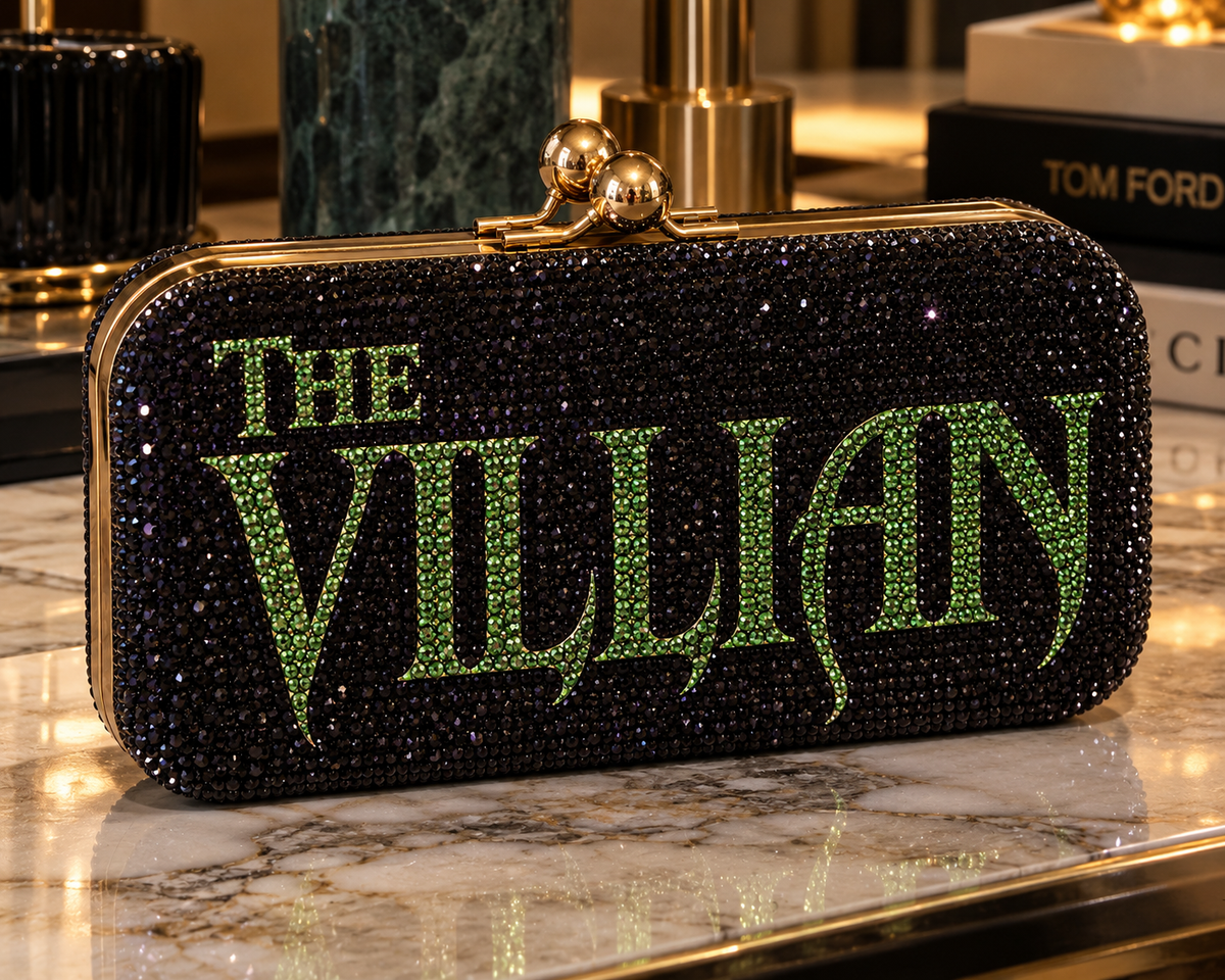 THE VILLAIN black crystal clutch with green rhinestone lettering on a marble surface